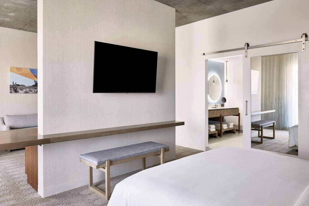 Ett modernt hotellrum med en stor säng, en platt-TV som är monterad på väggen, en bänk, en sminkbord med spegel och en dörr som leder till ett annat rum.
