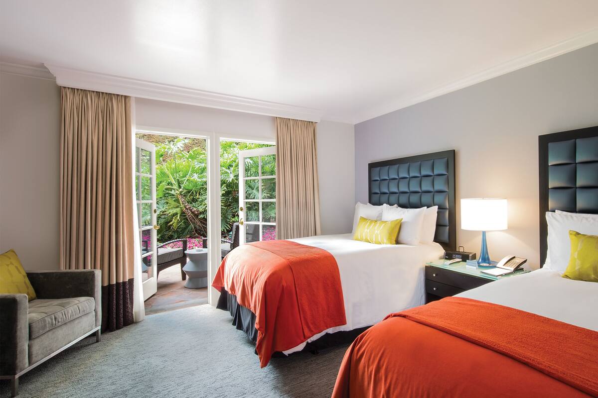 A hotel room with two beds, a sitting area with a chair and a table, and a view of a garden through a glass door.