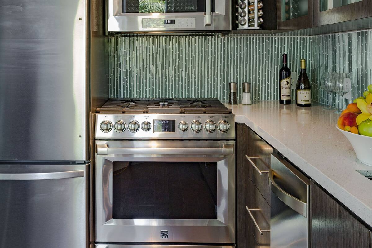 A modern kitchen with stainless steel appliances, a microwave, oven, and a gas stove. There are wine bottles and a fruit bowl on the counter.