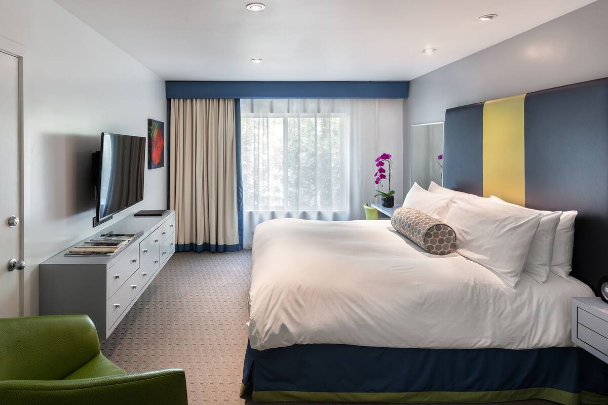 A modern bedroom with a large bed, a television on a dresser, a green chair, and a window with curtains.