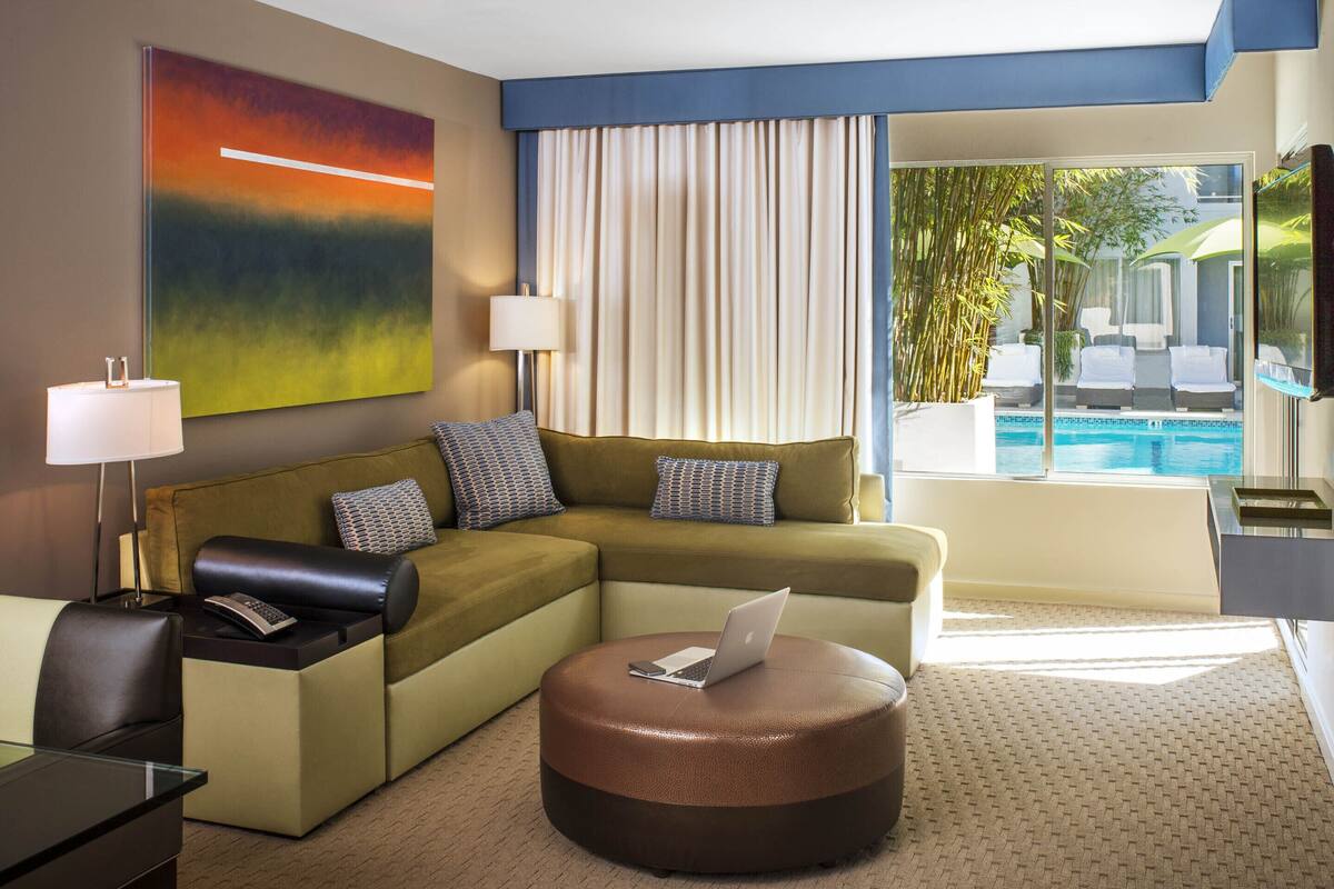 A modern hotel room with a sectional sofa, a round ottoman with a laptop, a desk with a lamp, and a view of a pool through large windows.