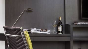 A modern office chair with a high backrest and a metallic frame, a desk with a laptop, a wine bottle, and a glass, and a wall-mounted shelf with decorative items.