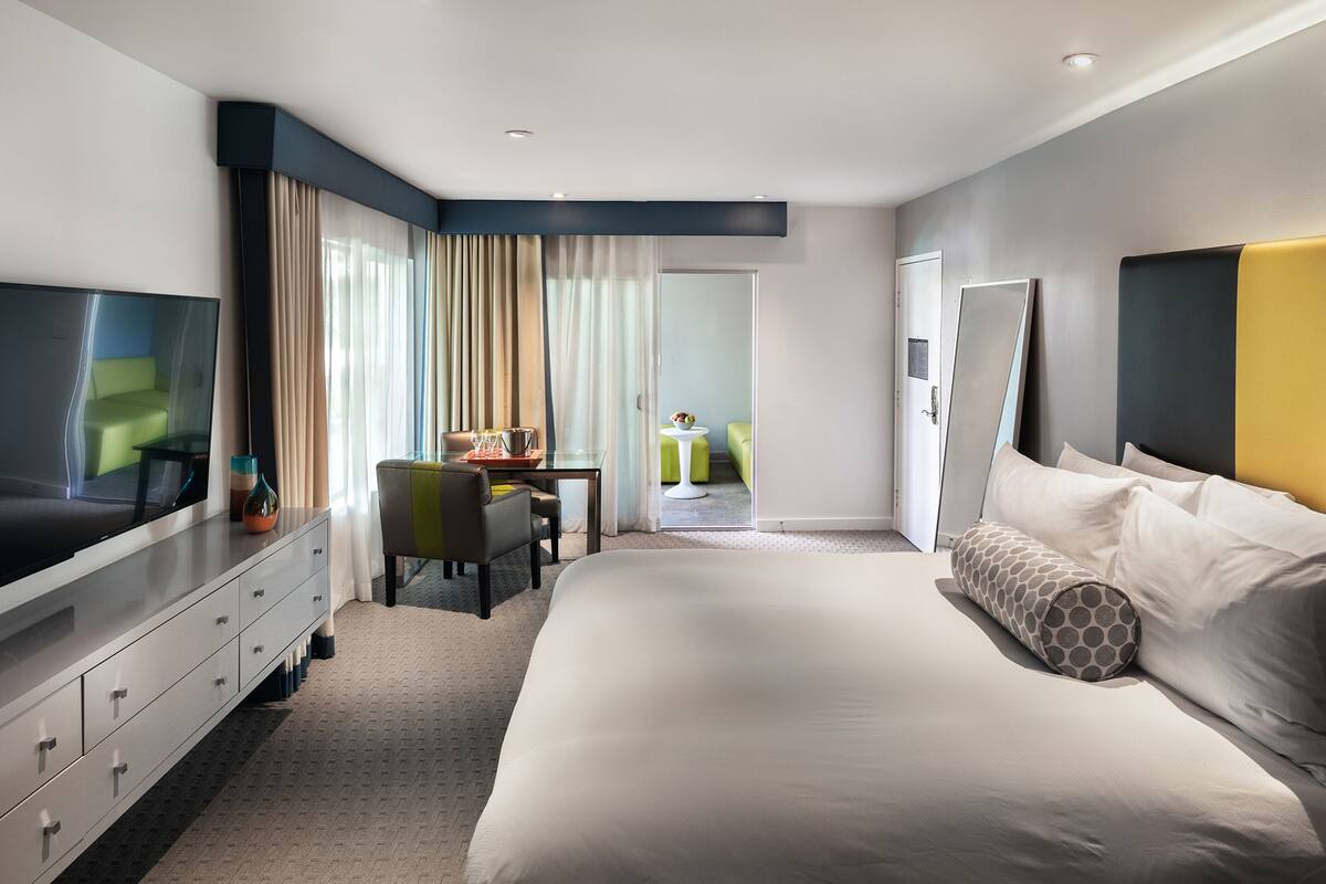 A modern hotel room with a large bed, a TV, a desk with a chair, and a small balcony.