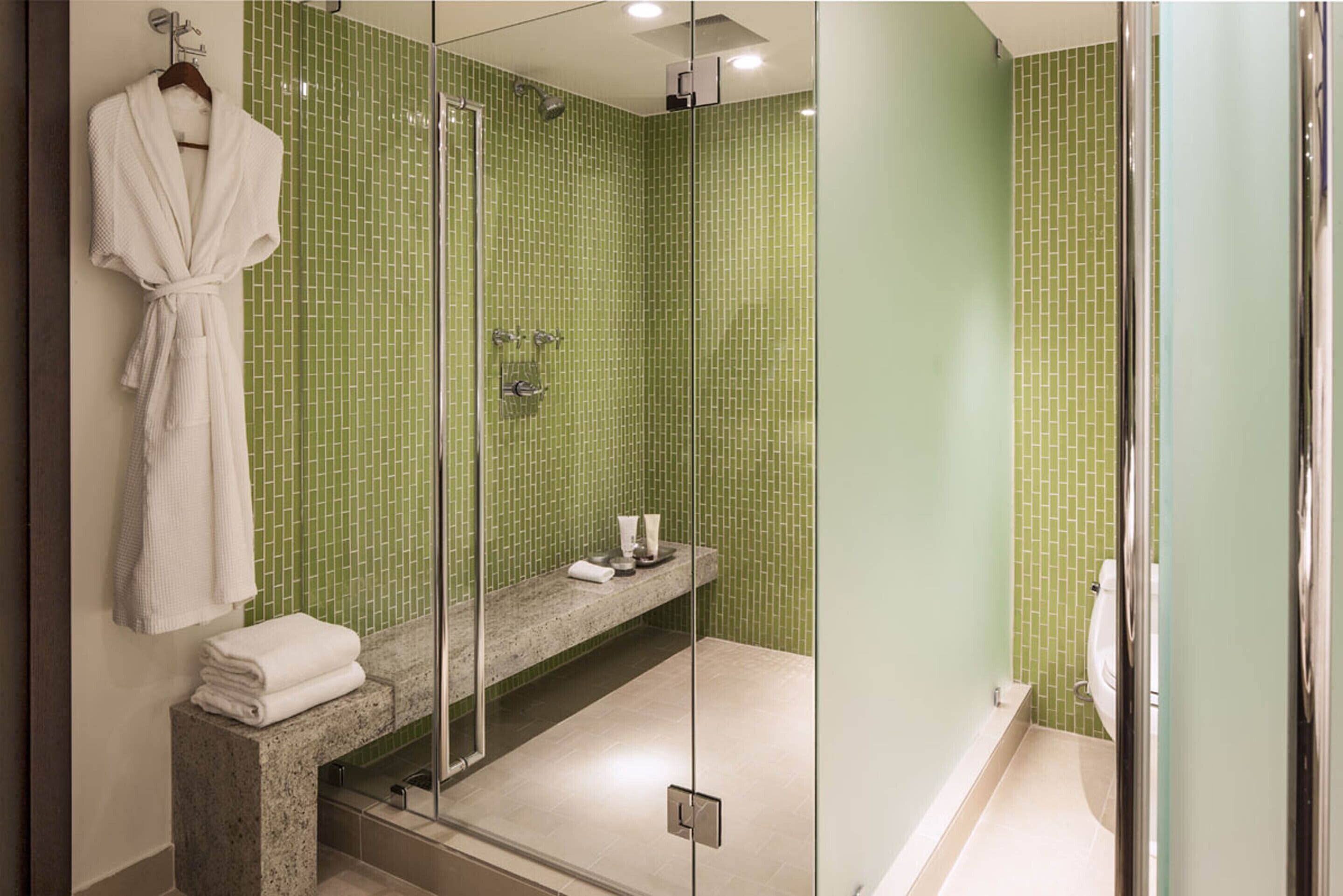 A bathroom with a glass shower enclosure, a bench with toiletries, and a hanging bathrobe.