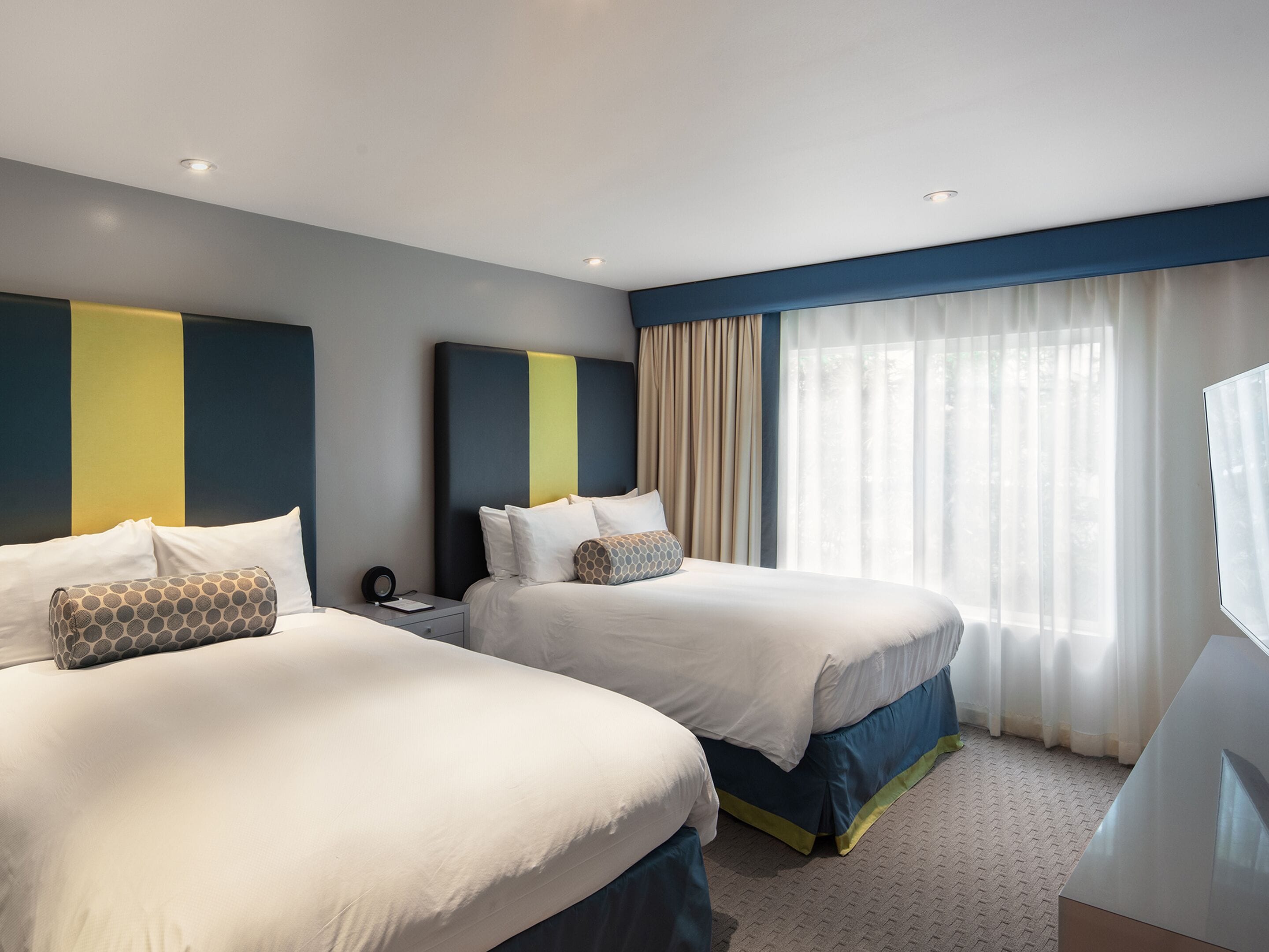 Une chambre d’hôtel avec deux lits, une table de chevet et une grande fenêtre avec des rideaux légers.