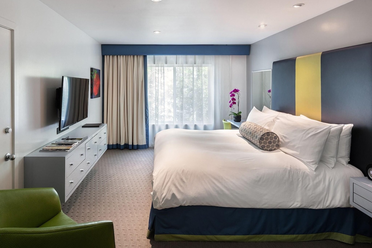 A modern hotel room with a bed, a television, a desk, and a green chair.