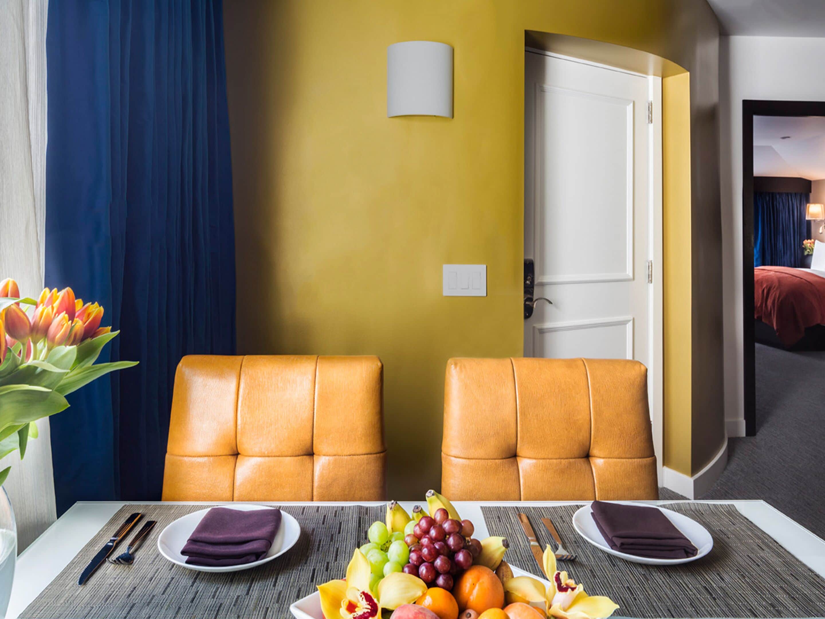 Habitación de hotel con dos sillones de cuero, una mesa de comedor con fruta y servilletas, una cama con una colcha roja y una pared amarilla con una lámpara.