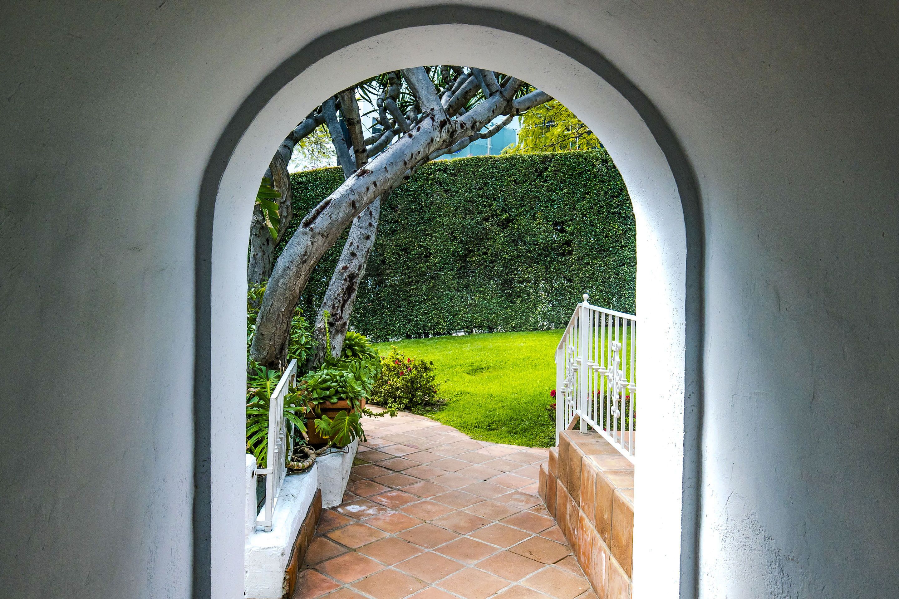 A view through an arched doorway leading to a paved area with a white railing and a green hedge in the background.