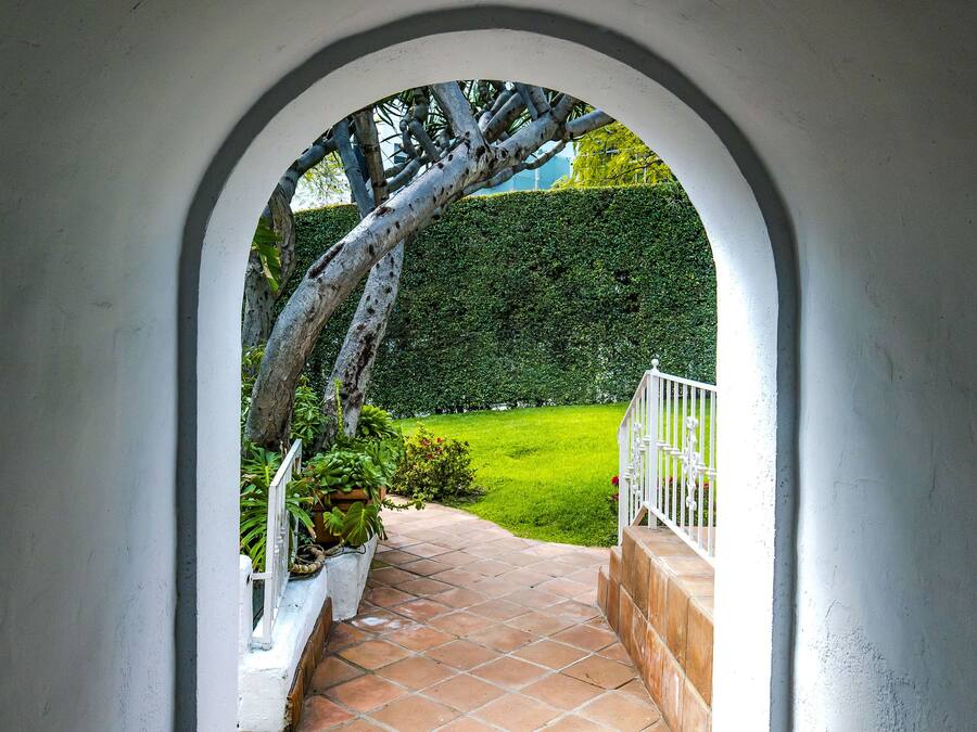 A view through an arched doorway leading to a paved area with a white railing and a green hedge in the background.