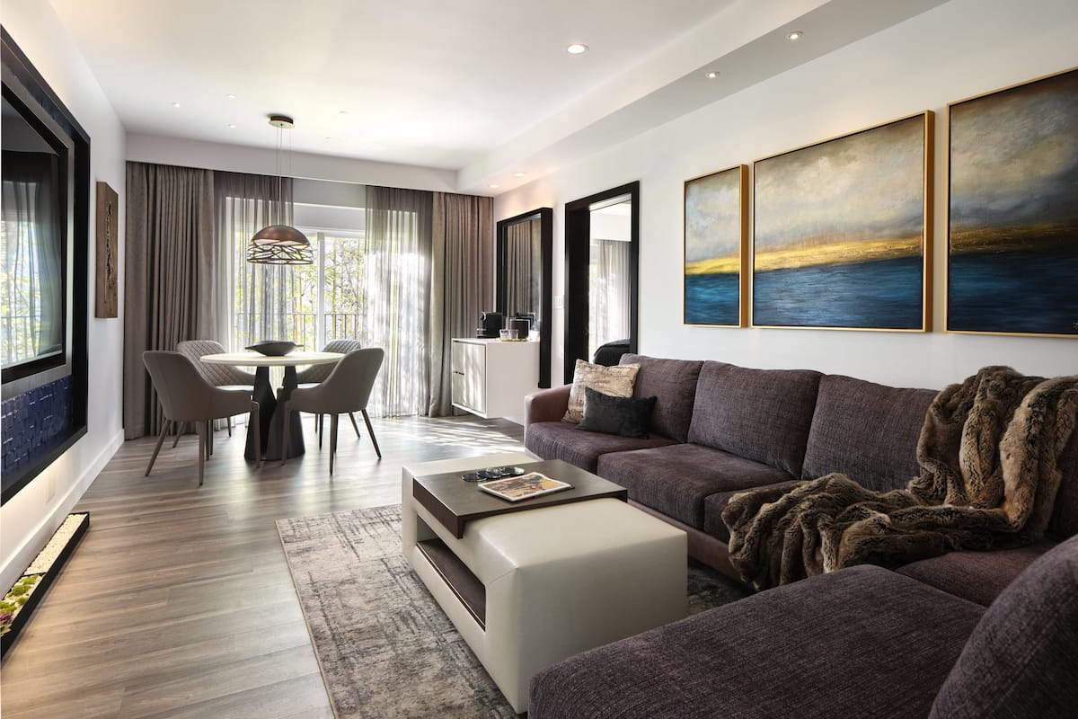 A modern living room with a sofa, coffee table, and dining area. There are three paintings on the wall and a large window with curtains.