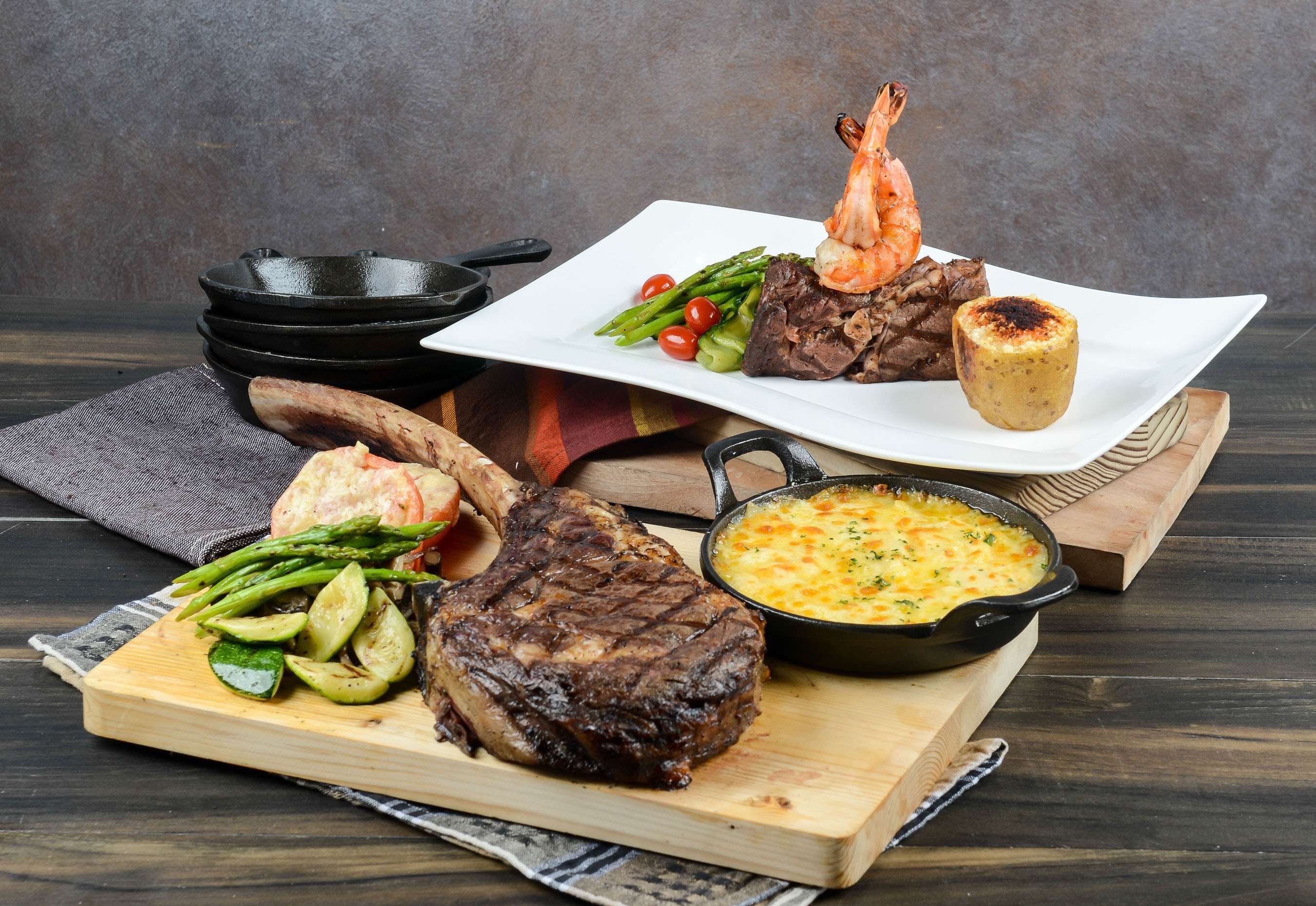 A variety of dishes including steak, shrimp, asparagus, and a baked potato on a wooden board.