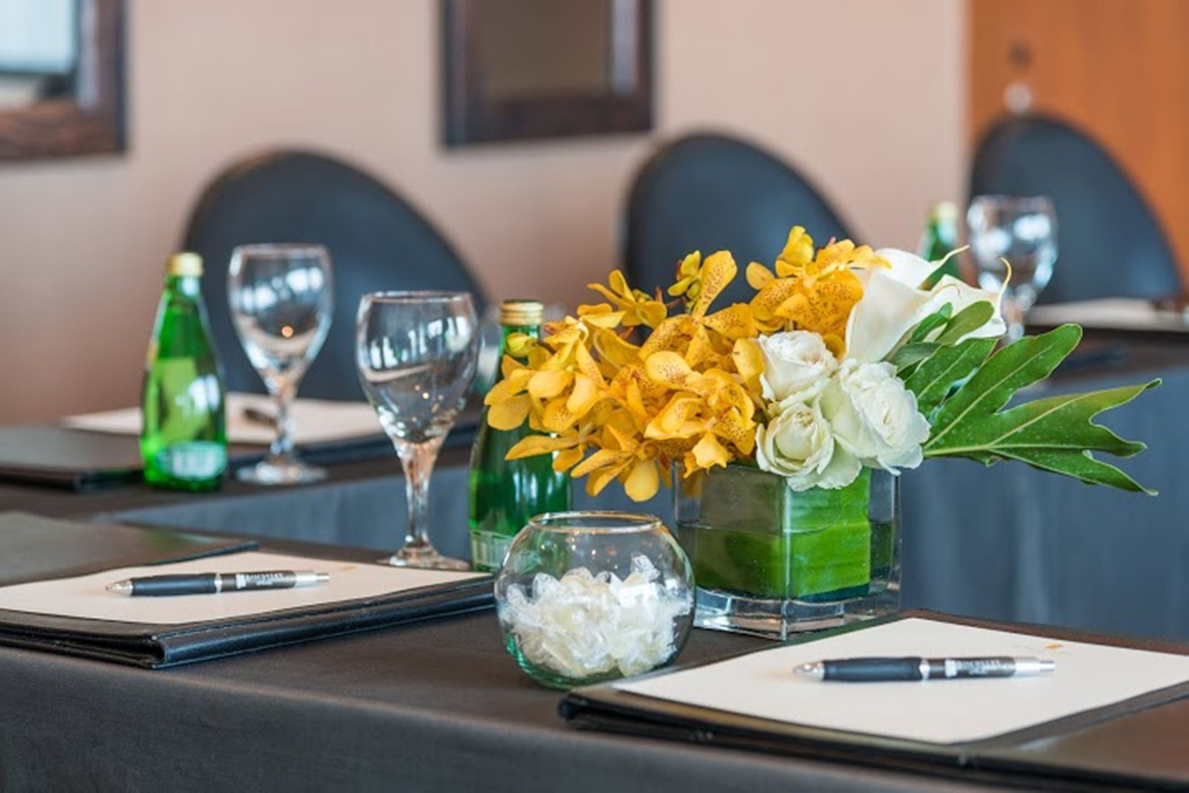 A conference table set with a pen, notepad, glass, and a vase with yellow and white flowers.
