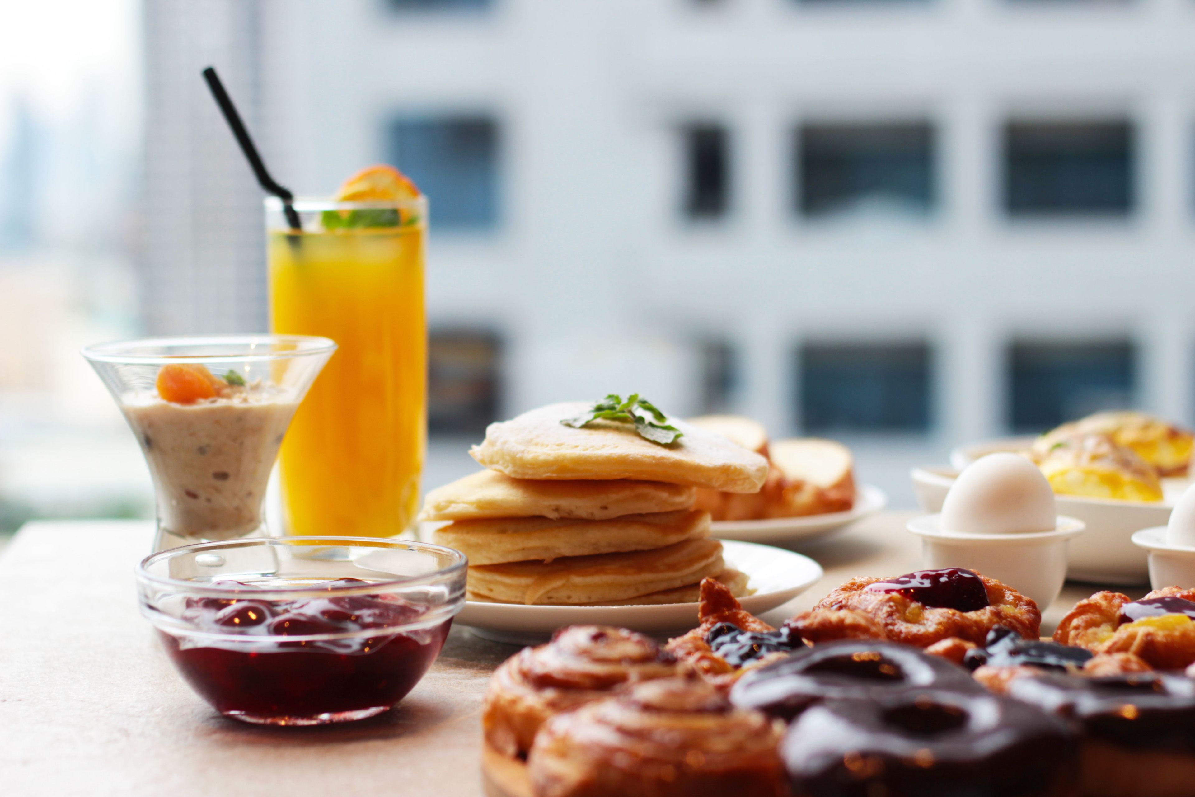 Una mesa de desayuno con tortitas, un vaso de zumo de naranja, un bol de mermelada y bollería.