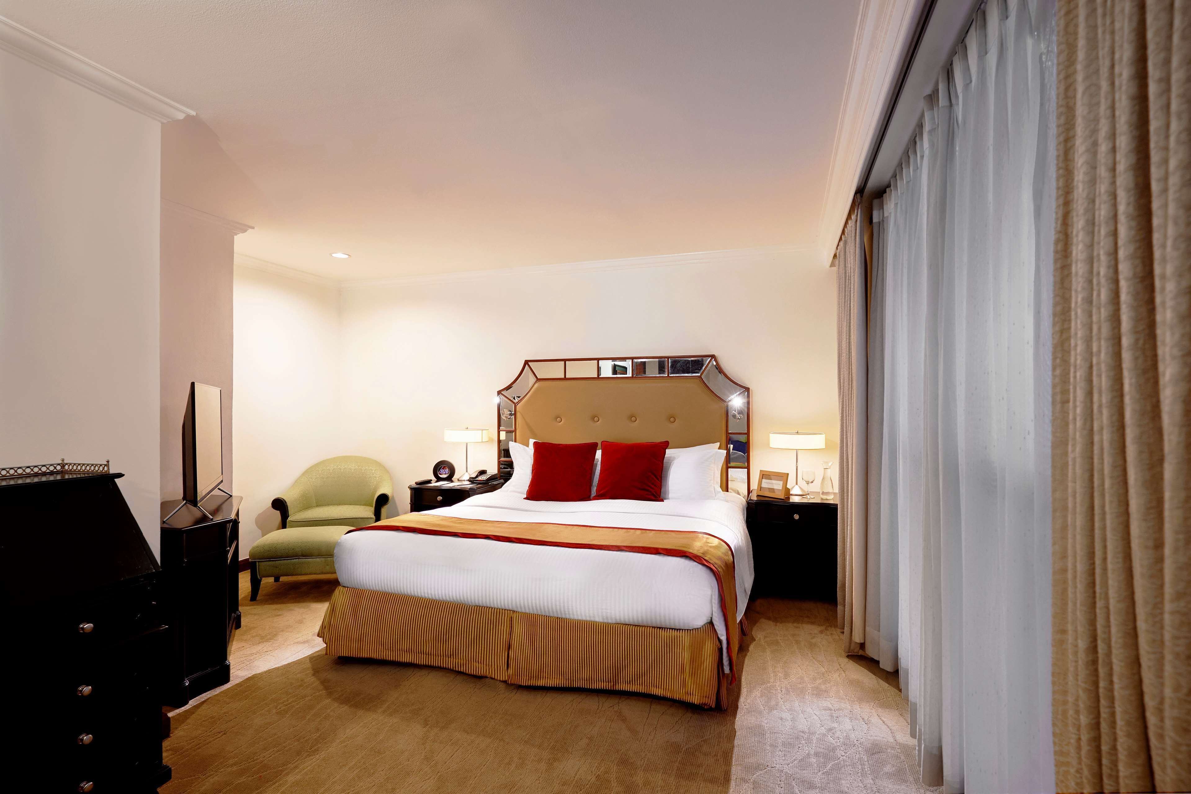 A hotel room with a large bed, a green armchair, a dresser, and a nightstand with a lamp.
