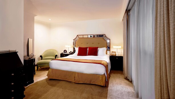 Quarto de hotel com uma cama grande, uma poltrona verde, um penteado e uma mesa de cabeceira com uma luminária.