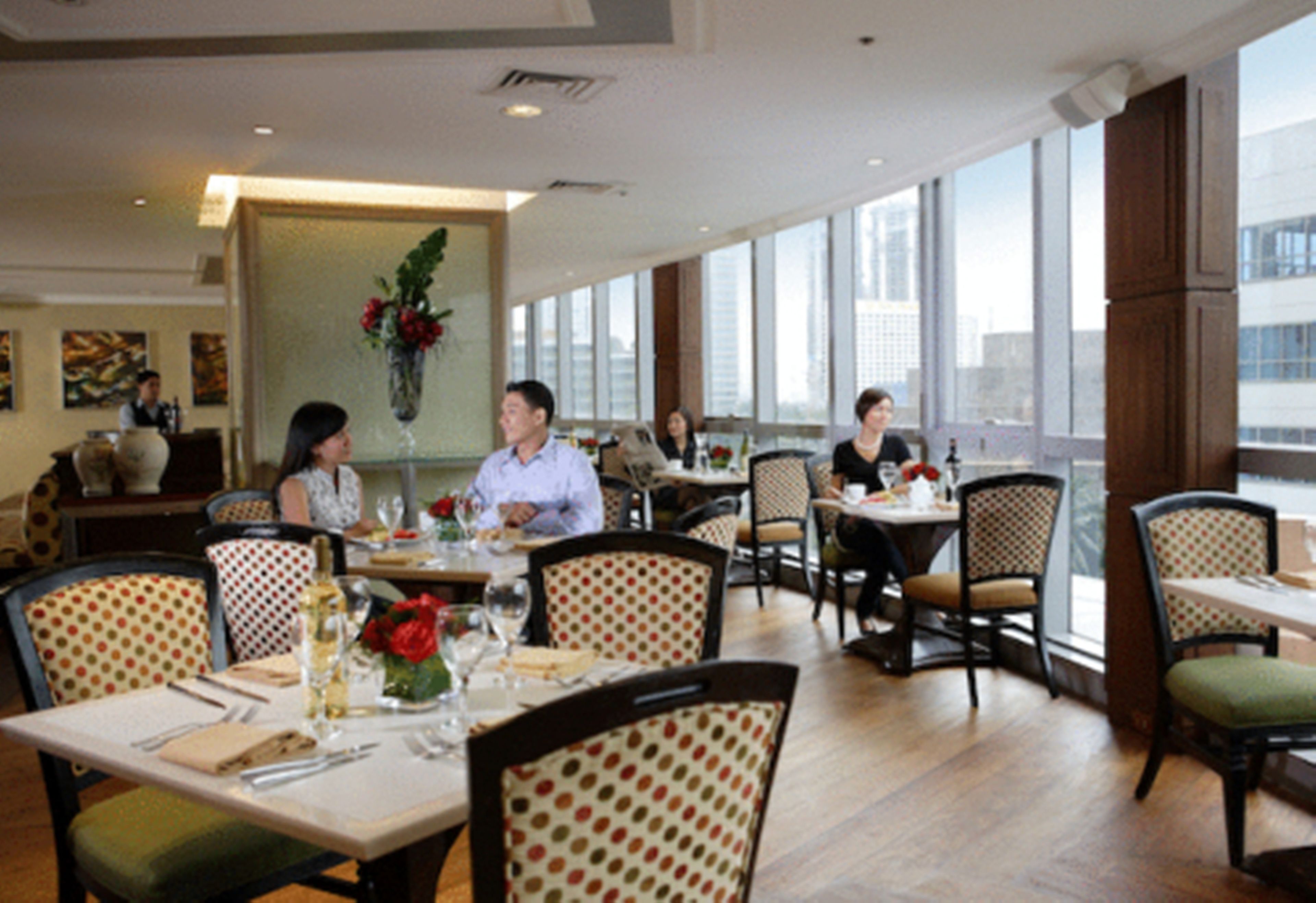 Un restaurante con techos altos, amplias ventanas y vistas a la ciudad. Las mesas están dispuestas con cubiertos y servilletas.