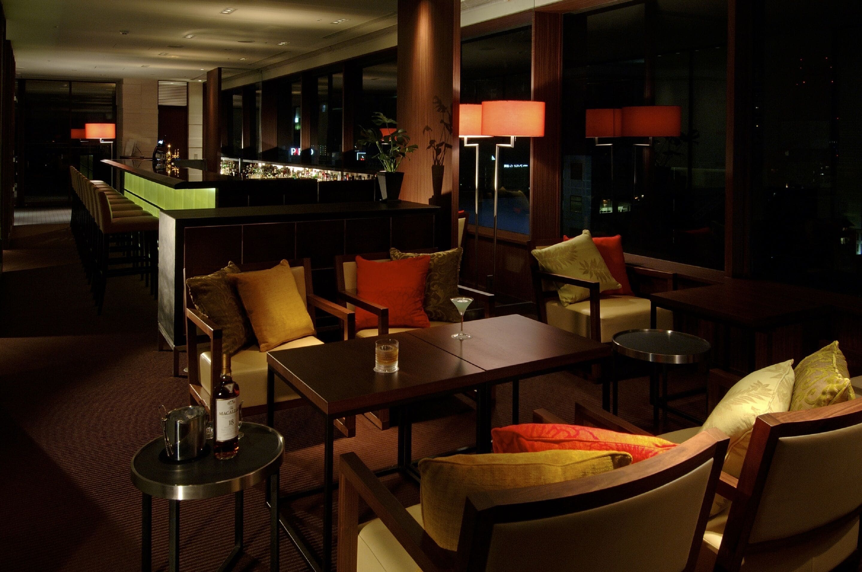 A dimly lit hotel lobby with a bar area, seating arrangements, and large windows.