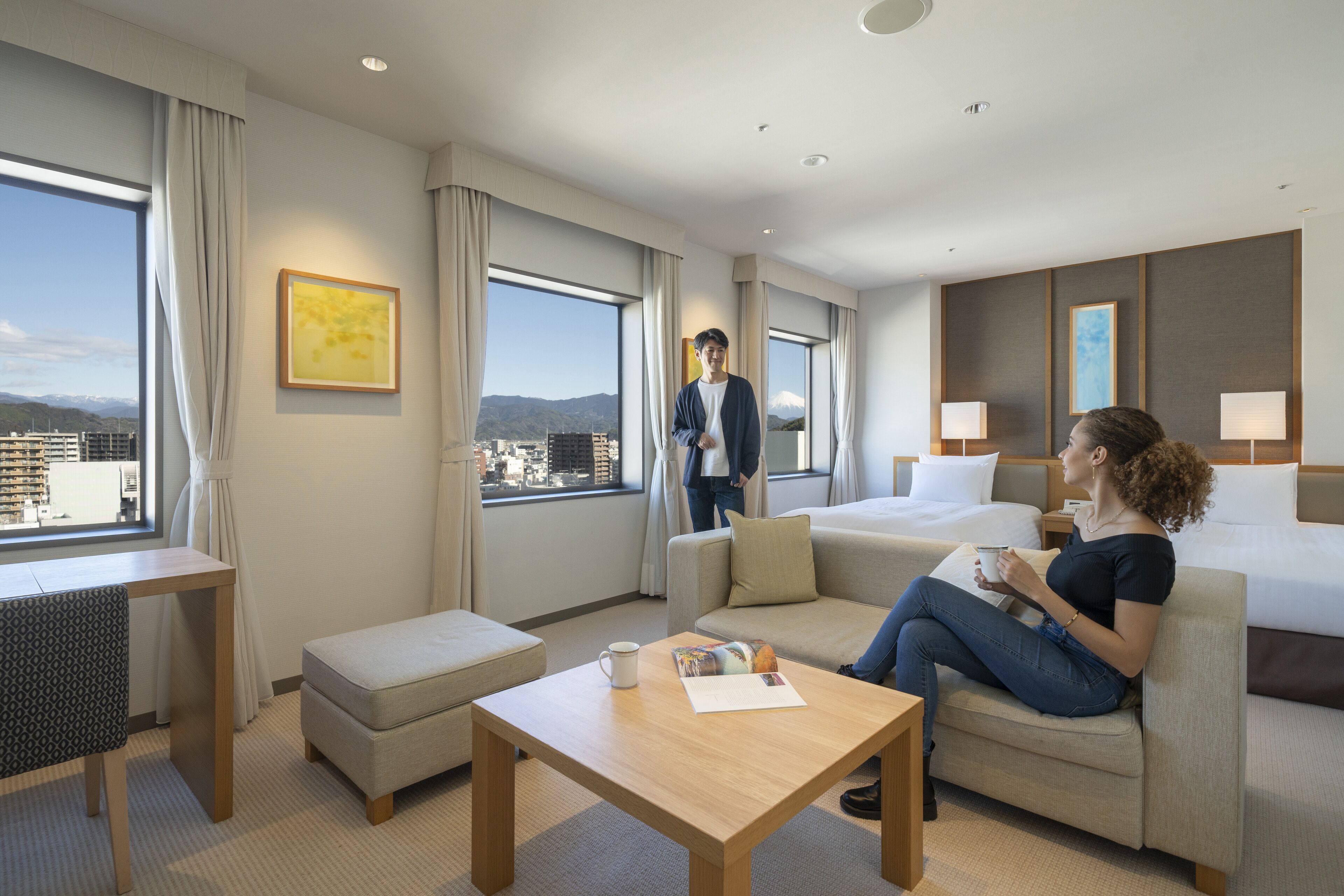 A modern hotel room with a large window, a sofa, a coffee table, and a desk.