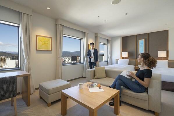 A modern hotel room with a large window, a sofa, a coffee table, and a desk.