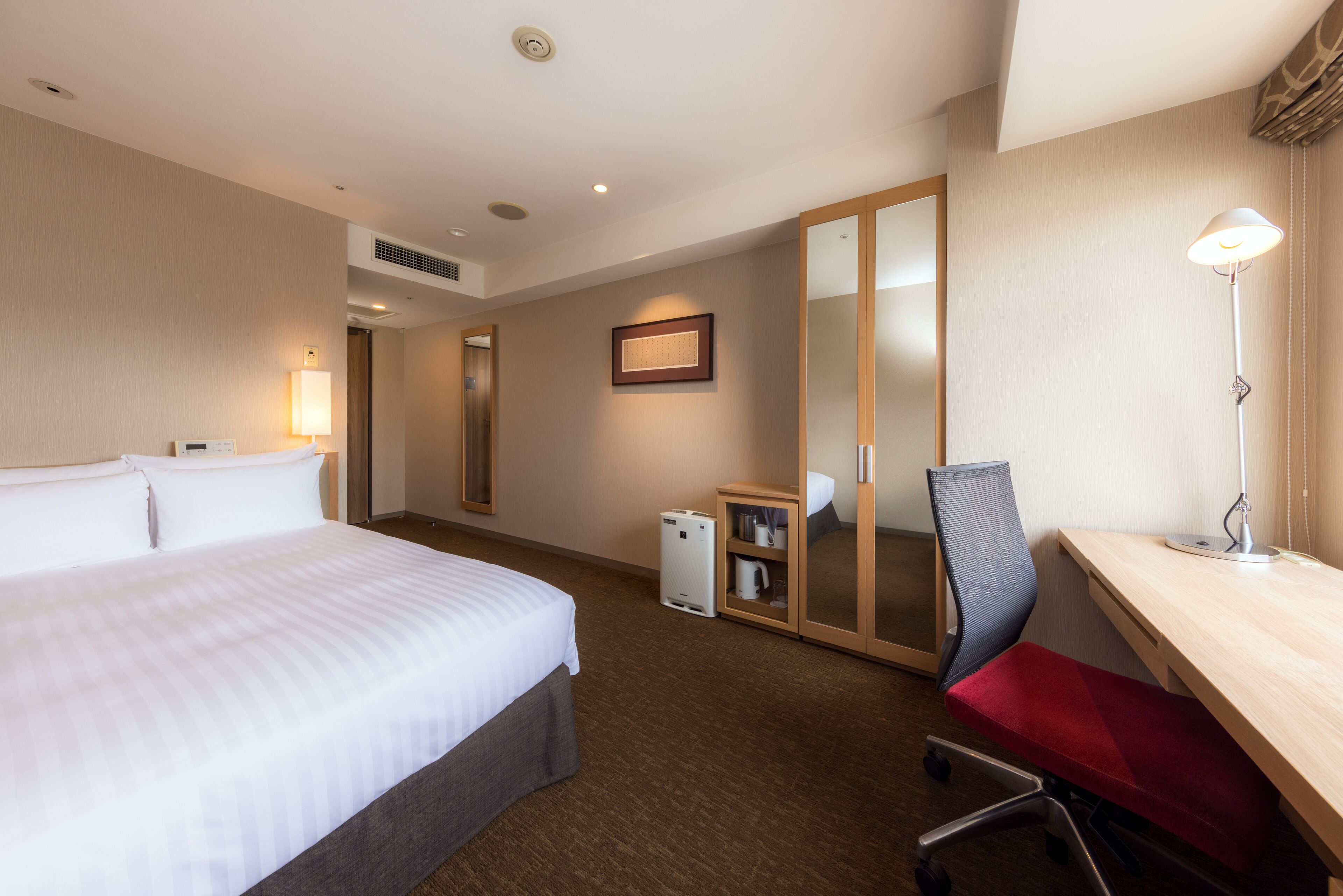A hotel room with a bed, desk, chair, lamp, and a mirror.