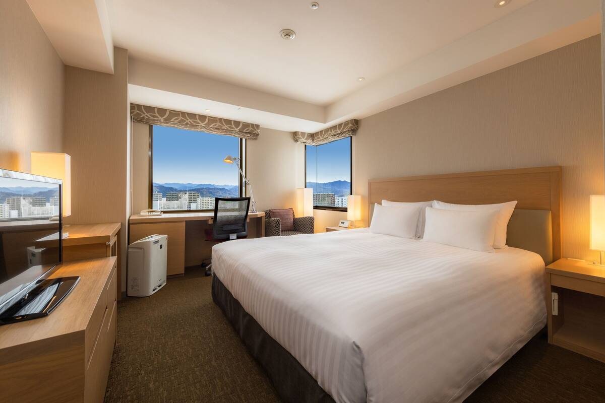Une chambre d’hôtel avec un grand lit, un bureau équipé d’un ordinateur, une chaise et une vue sur le paysage urbain par les fenêtres.