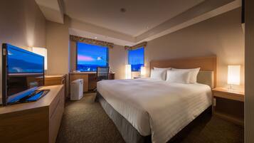 A hotel room with a large bed, a desk with a computer, a TV, and a view of the cityscape at dusk.