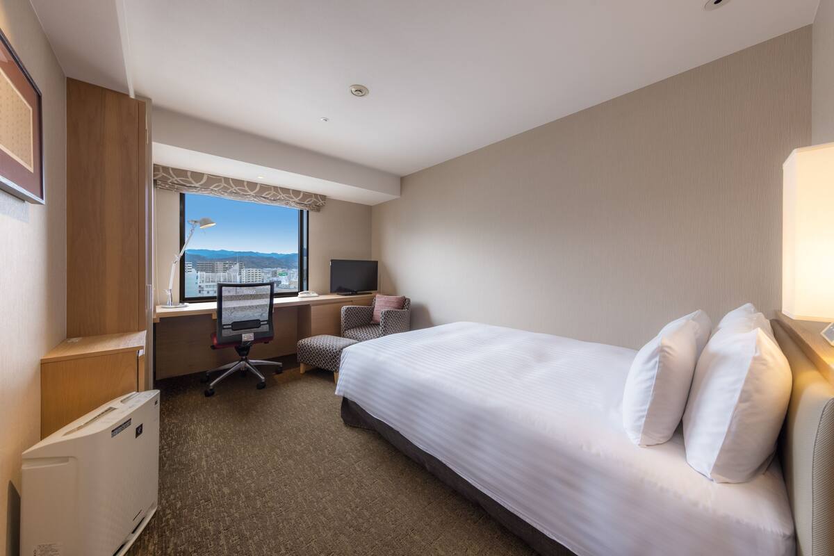 Une chambre d’hôtel avec un lit, un bureau, une chaise et une vue sur le paysage urbain par la fenêtre.