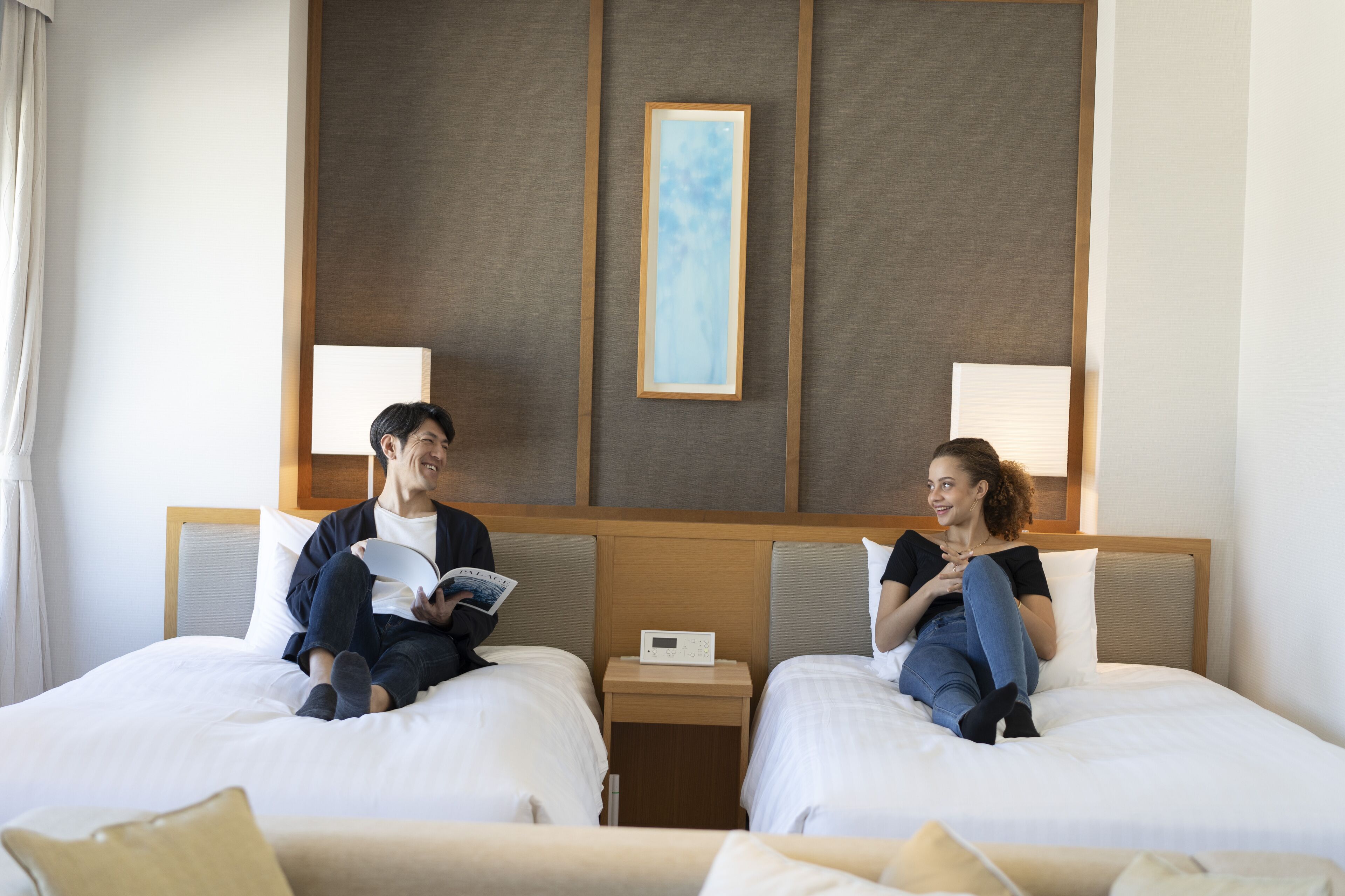 Two beds in a hotel room with a bedside table between them, a framed picture on the wall, and a window with curtains.