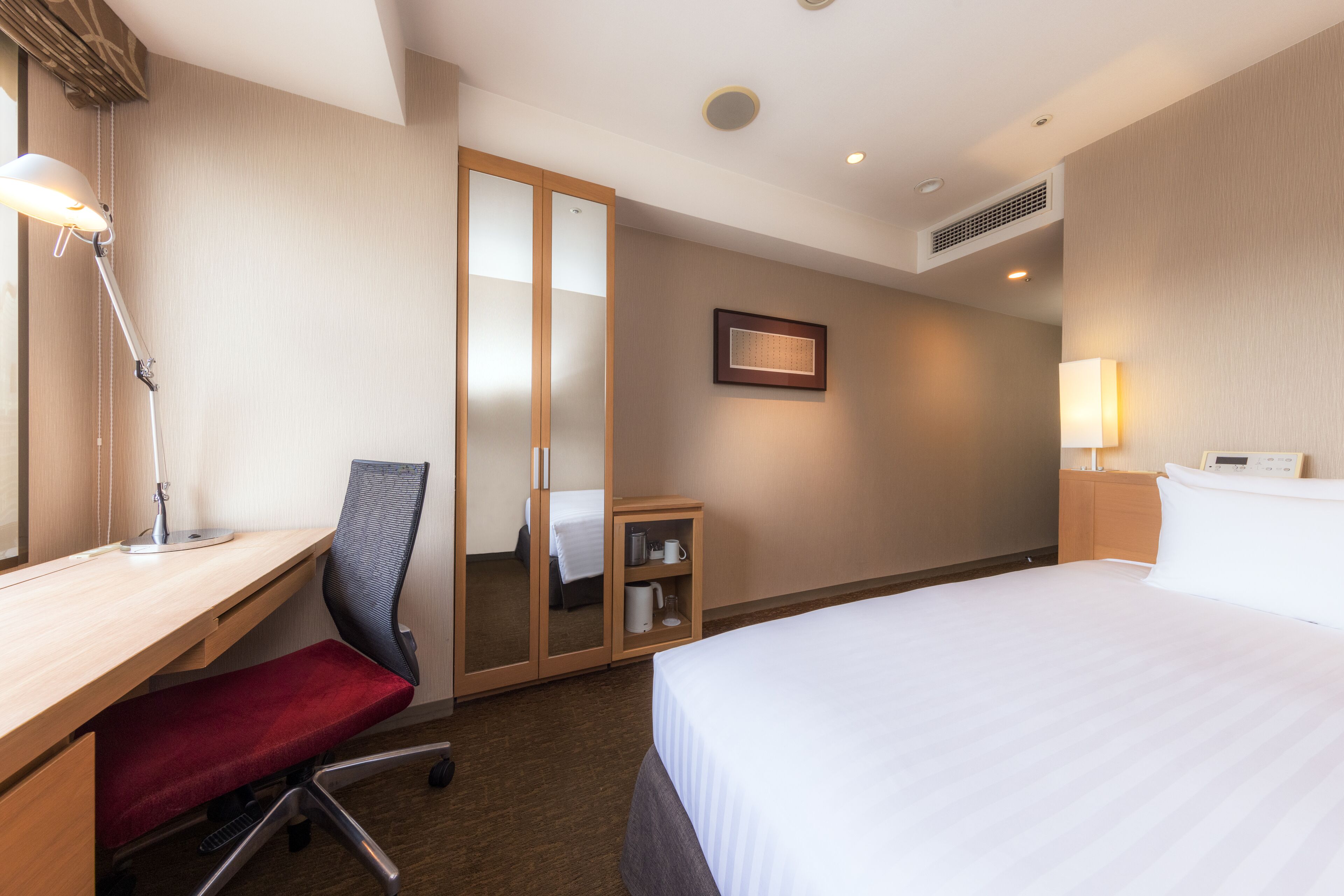 A hotel room with a bed, desk, chair, mirror, bedside table, and a small cabinet.