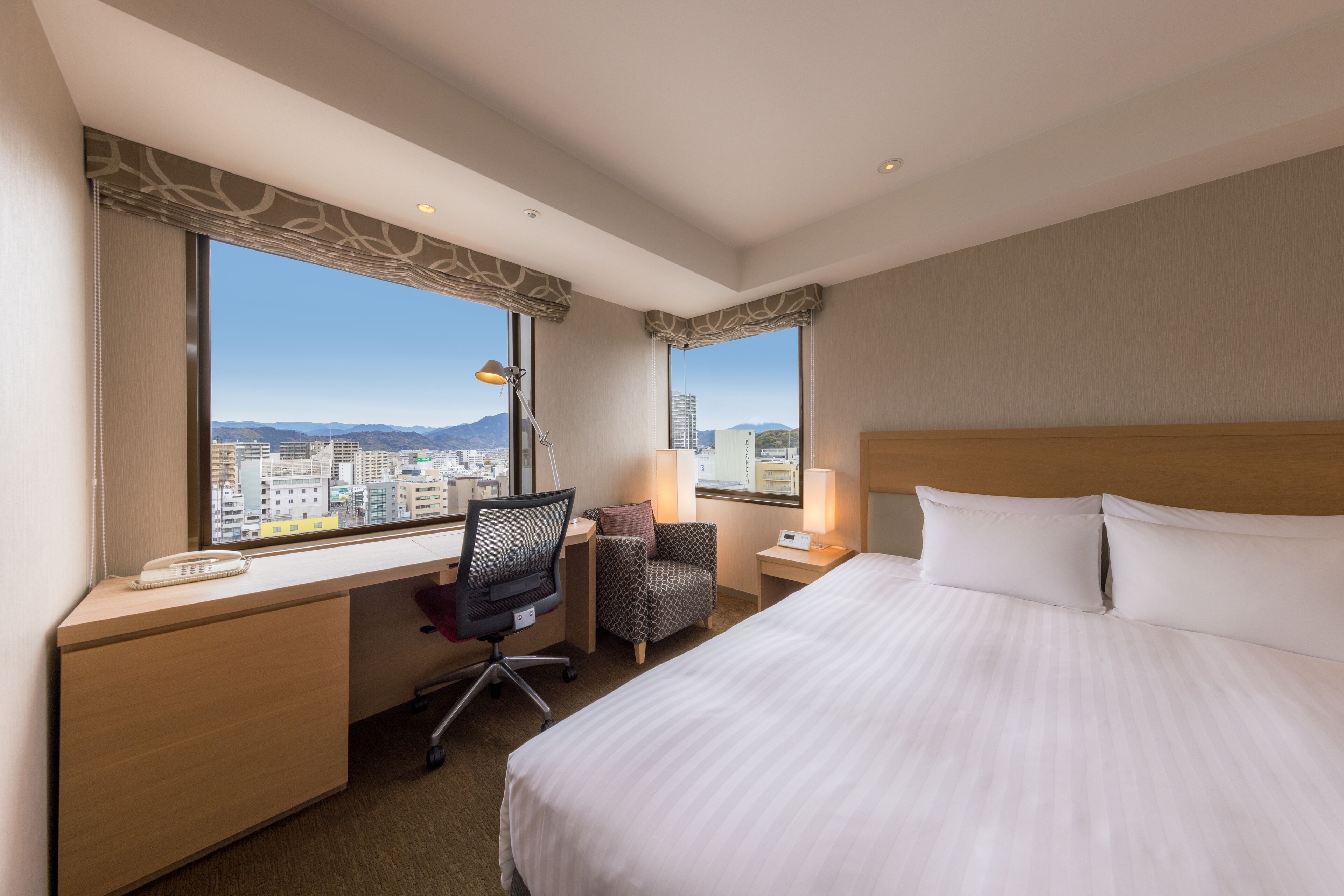 A hotel room with a large bed, a desk with a chair, a lamp, and a view of the city through large windows.