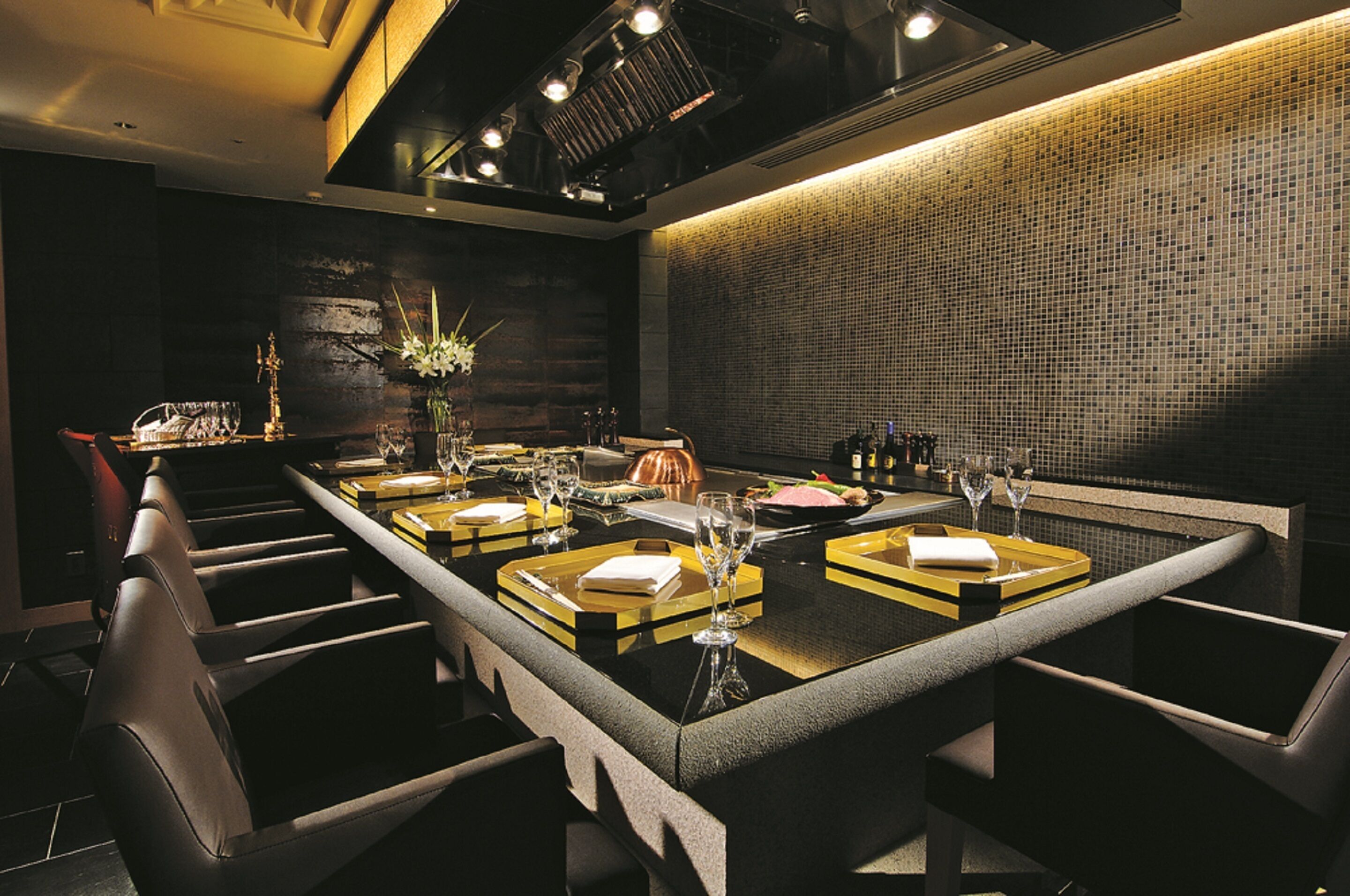 A modern dining area with a long table set for a meal, featuring a central floral arrangement, wine glasses, and a variety of dishes.