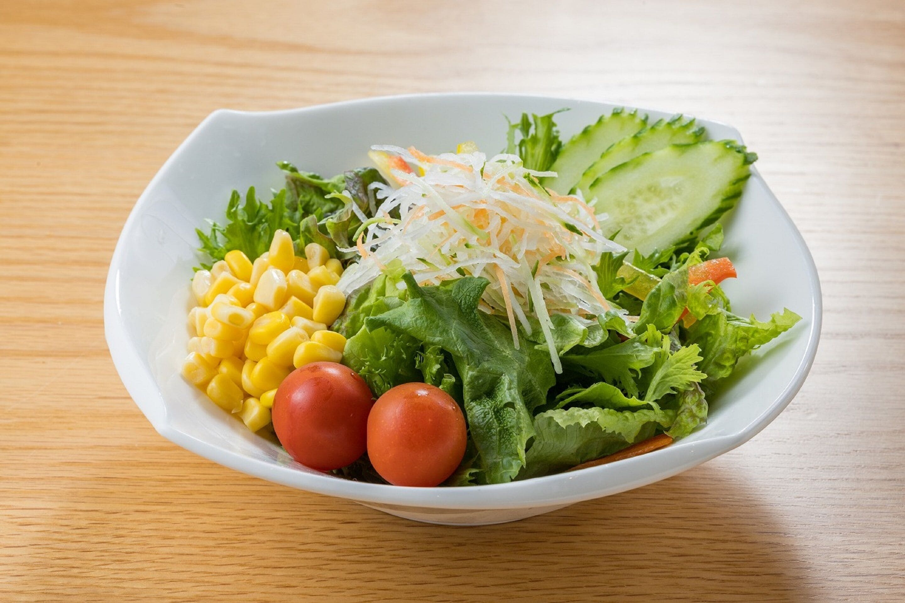 A white bowl containing a salad with lettuce, cucumber, shredded carrots, corn, and tomatoes.