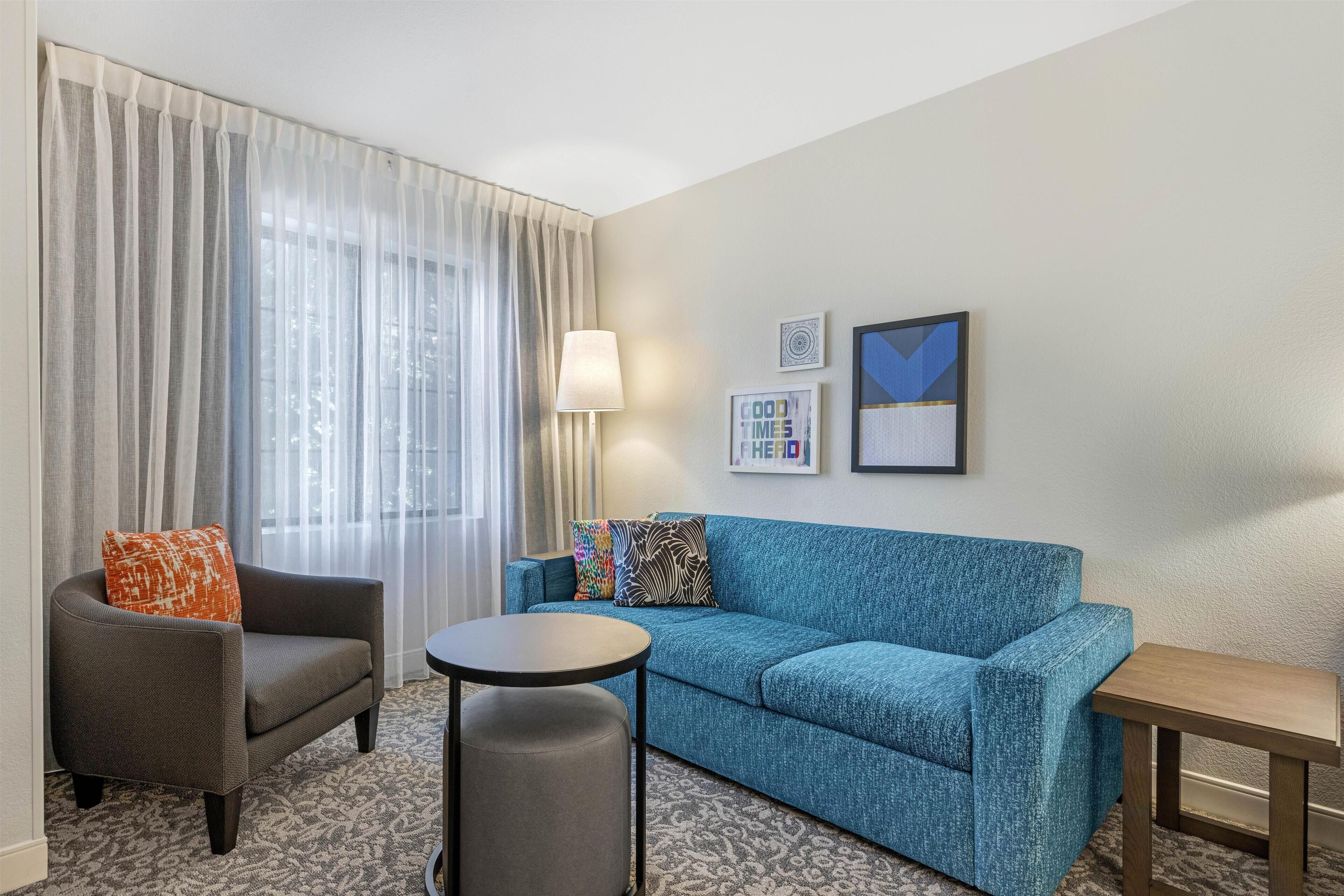A hotel room with a grey armchair, a blue sofa, a small round table, and a lamp. There are two framed pictures on the wall.