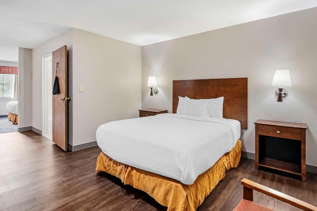 Quarto de hotel com uma cama grande, duas mesas de cabeceira, uma cabeceira de madeira e uma porta que dá para outro quarto.