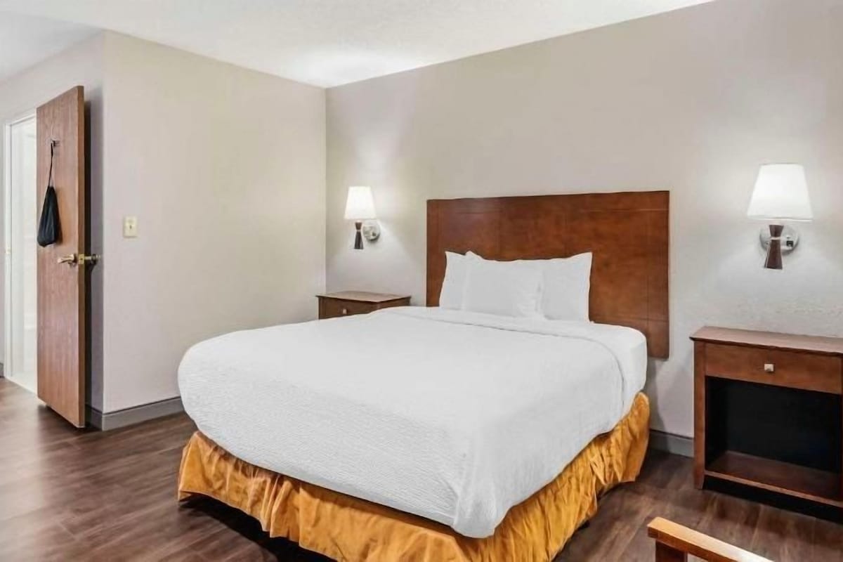 Quarto de hotel com uma cama grande, duas mesas de cabeceira com abajures, um painel de madeira na cabeceira e uma porta que dá para outro quarto.