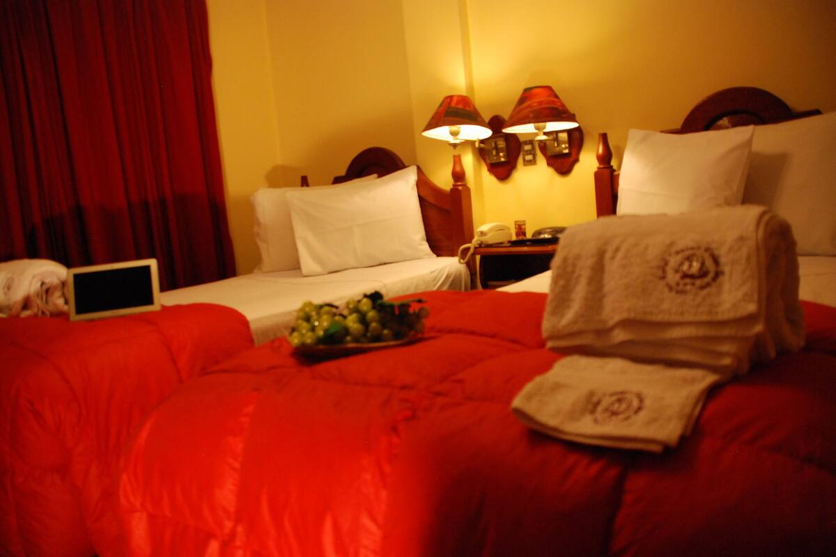 A hotel room with a red bedspread, a laptop on the left side, a fruit bowl on the bed, and a towel on the right side.