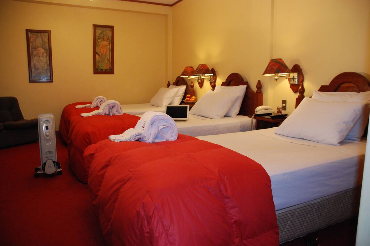 A hotel room with two beds, each with red bedding and white pillows. There is a desk with a laptop and a telephone, and a vacuum cleaner on the floor.