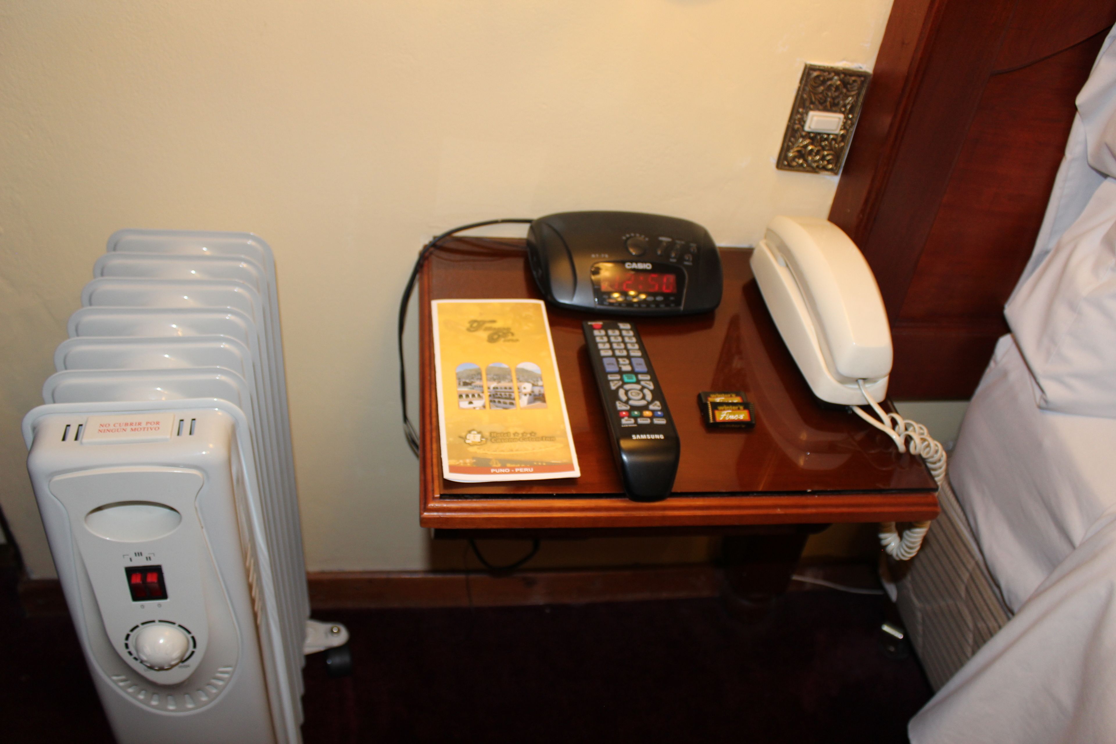 Habitación de hotel con panel de control, control remoto, teléfono y un folleto sobre la mesita de noche.