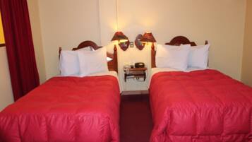 A hotel room with two single beds, each with a red bedspread and white pillows. There are two wall-mounted lamps on either side of the bed.