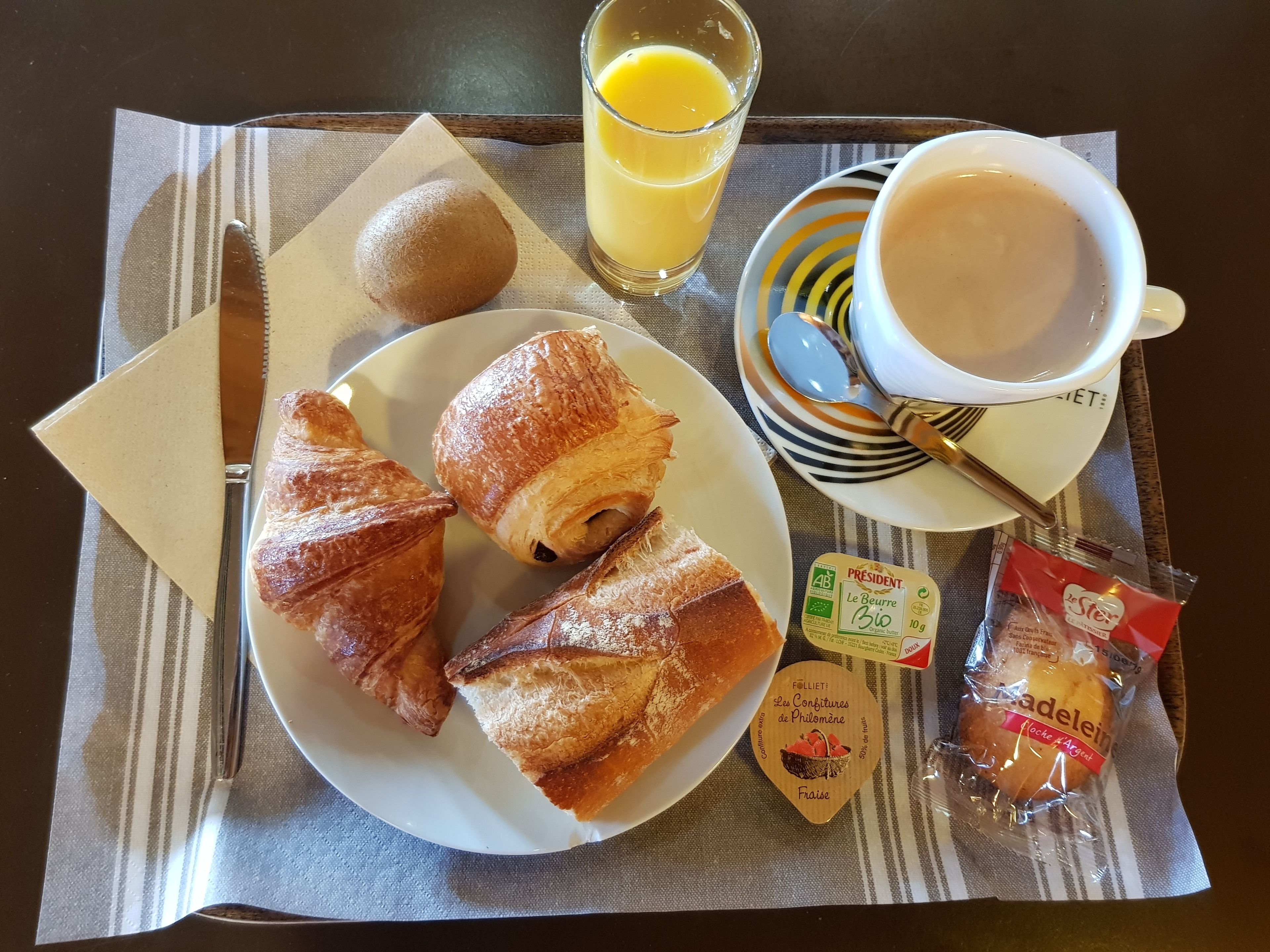 Un plateau de petit-déjeuner avec des croissants, un kiwi, un verre de jus d’orange, une tasse de café et des en-cas emballés.