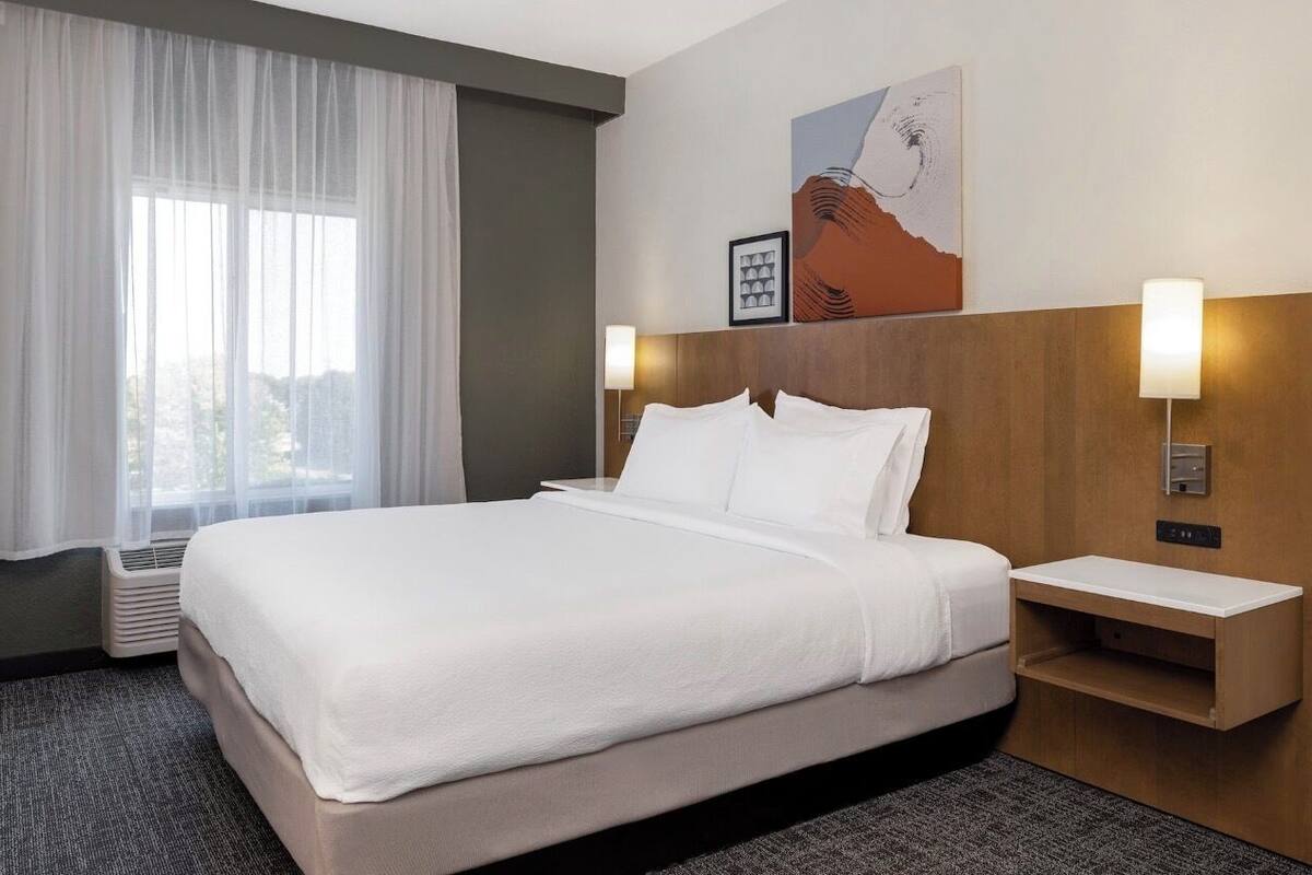 Une chambre d’hôtel avec un grand lit, du linge de lit blanc, une tête de lit en bois, deux tables de chevet avec des lampes, un radiateur fixé au mur et une fenêtre avec des rideaux légers.