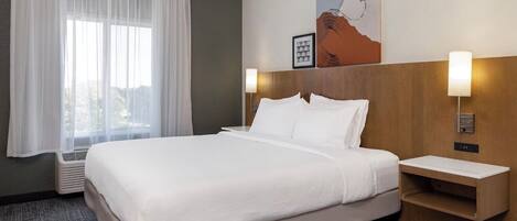A hotel room with a large bed, white linens, a wooden headboard, two bedside tables with lamps, a wall-mounted heater, and a window with sheer curtains.