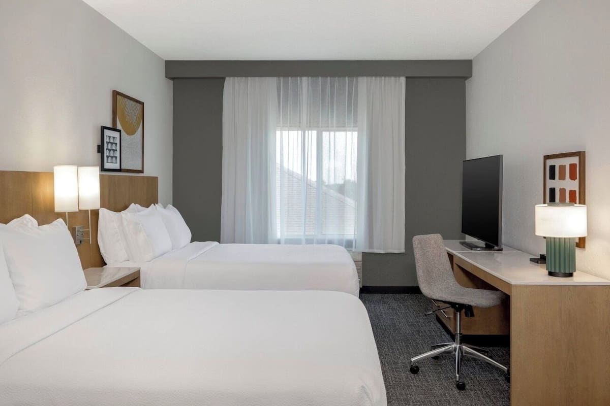Une chambre d’hôtel avec deux lits, un bureau, une chaise, une télévision et une fenêtre avec des rideaux.