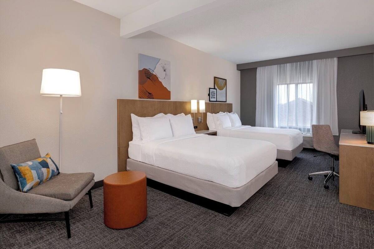 Une chambre d’hôtel avec deux lits, un bureau, une chaise, une lampe et des œuvres d’art accrochées au mur.