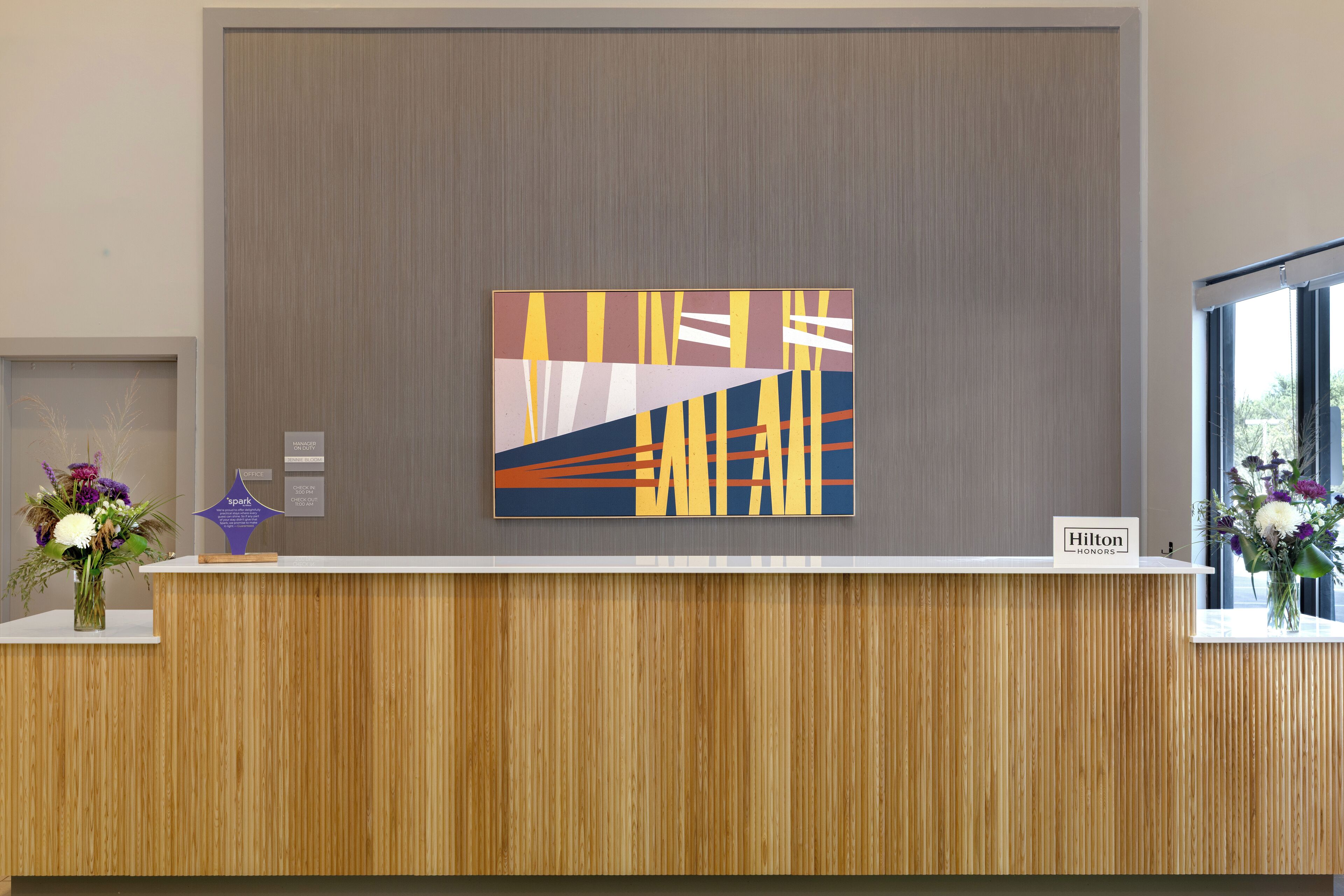 Un mostrador de recepción de hotel con diseño moderno, una gran obra de arte abstracta sobre él y un cartel que indica 'Hilton Honors'.