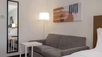 Un dormitorio con un sofá gris, una mesita blanca pequeña, una lámpara de pie, un espejo grande y un cuadro en la pared.
