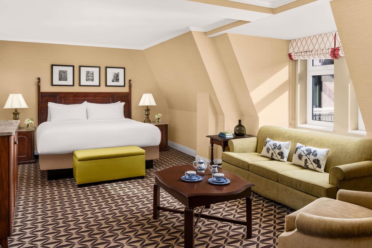 A hotel room with a bed, a sofa, a coffee table, a nightstand, and a window with a view.