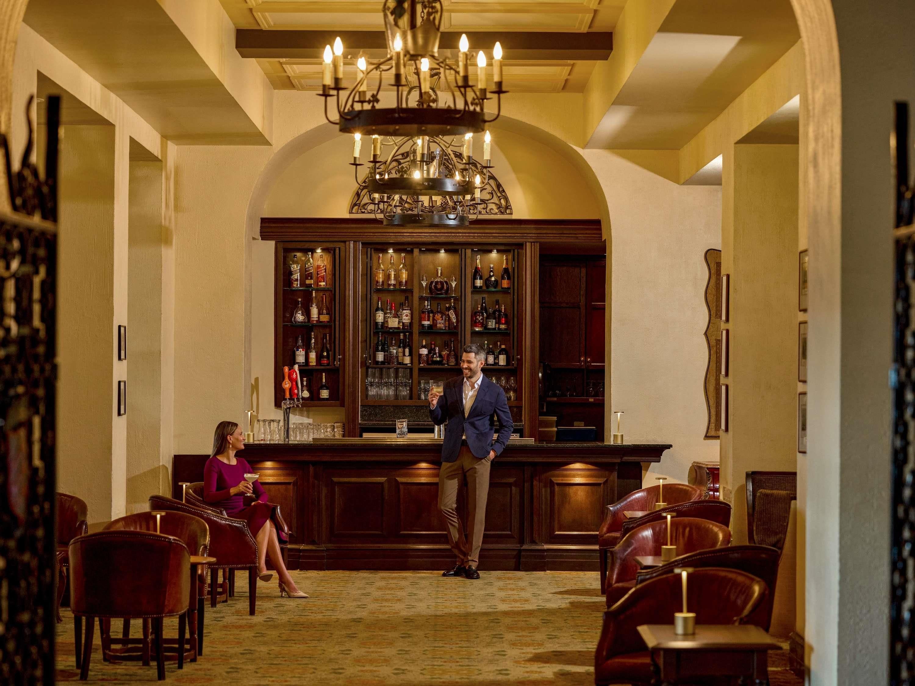 A bar area with a chandelier, wooden cabinets with liquor bottles, and leather chairs.
