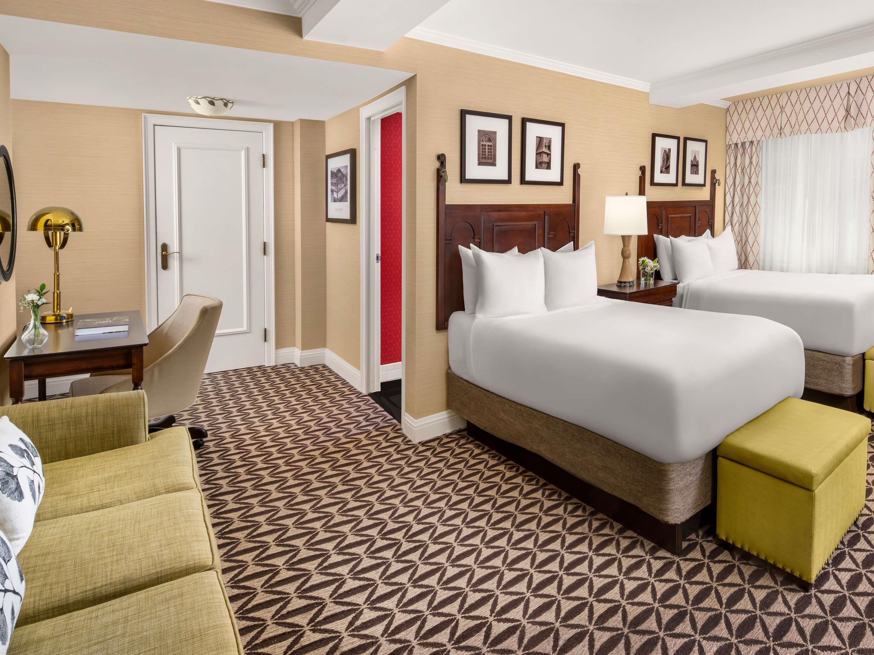 Ein Hotelzimmer mit einem großen Bett, zwei Einzelbetten, einem Schreibtisch mit Lampe, einem Sessel und einem gemusterten Teppich.