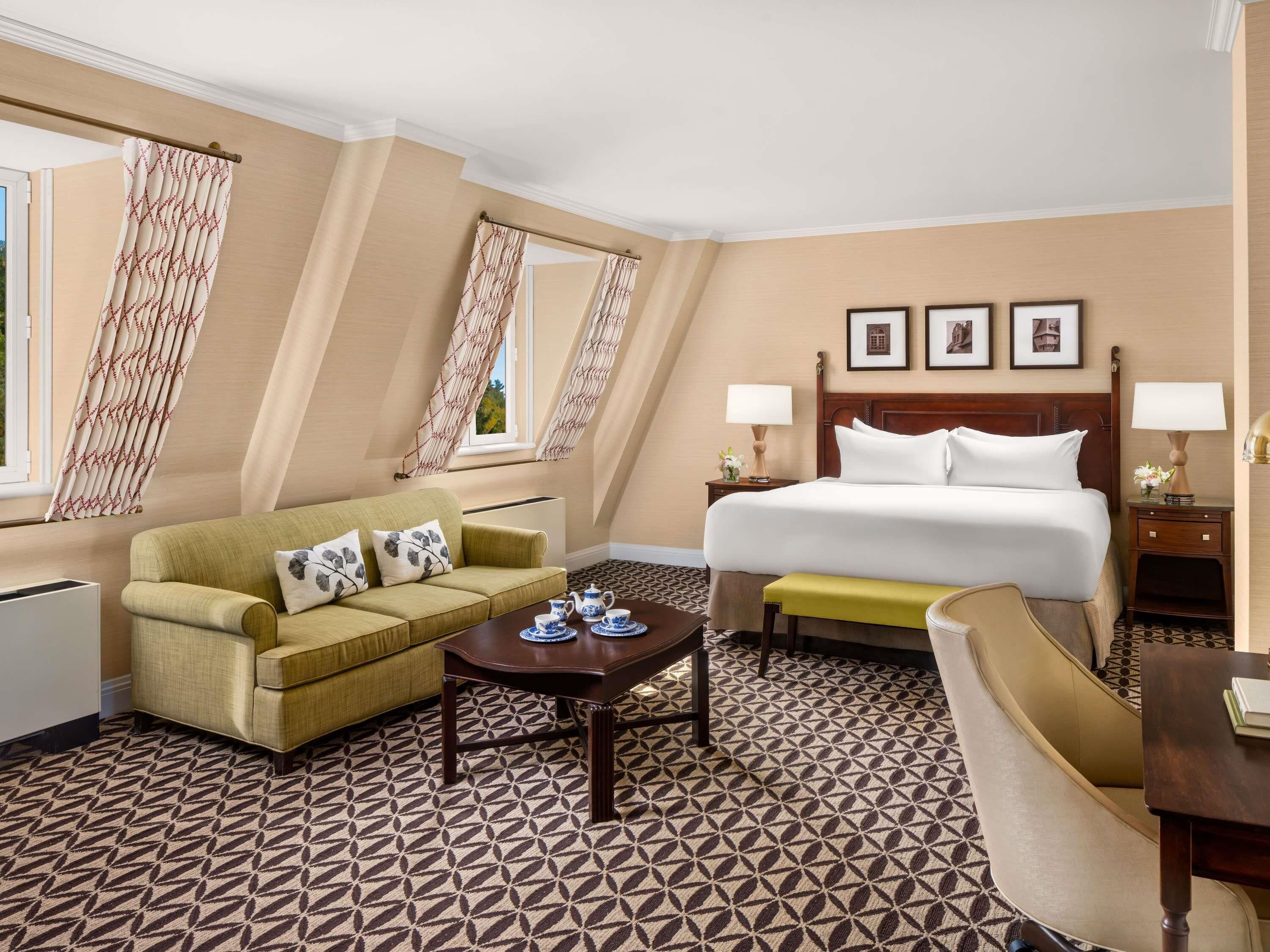 A hotel room with a bed, a sofa, a small table, and a chair. There are two bedside tables with lamps. The room has a patterned floor and a large window with curtains.